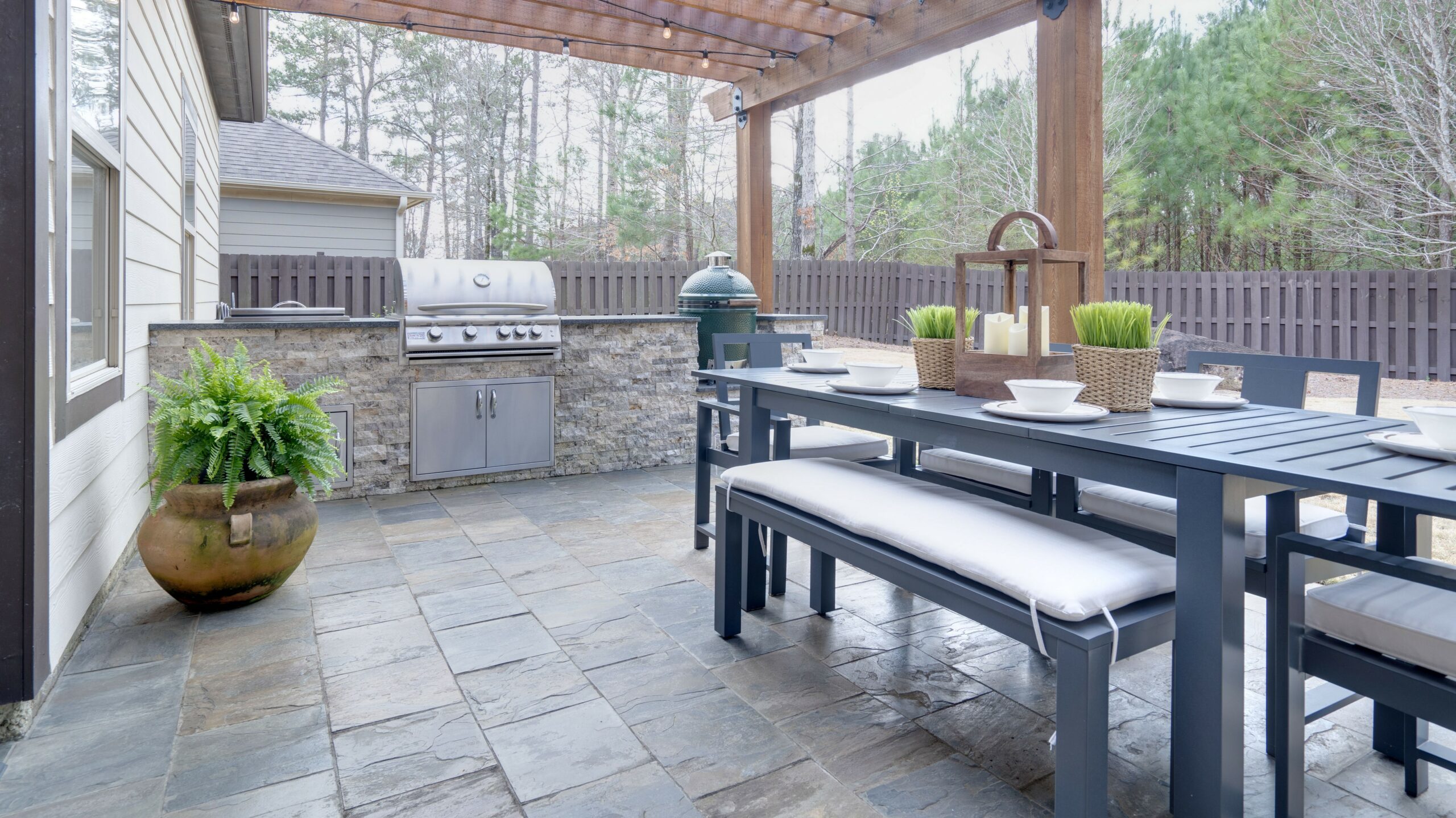 Pretty summer outdoor kitchen with table set and grilling station underneath wooden arbor on stone patio.